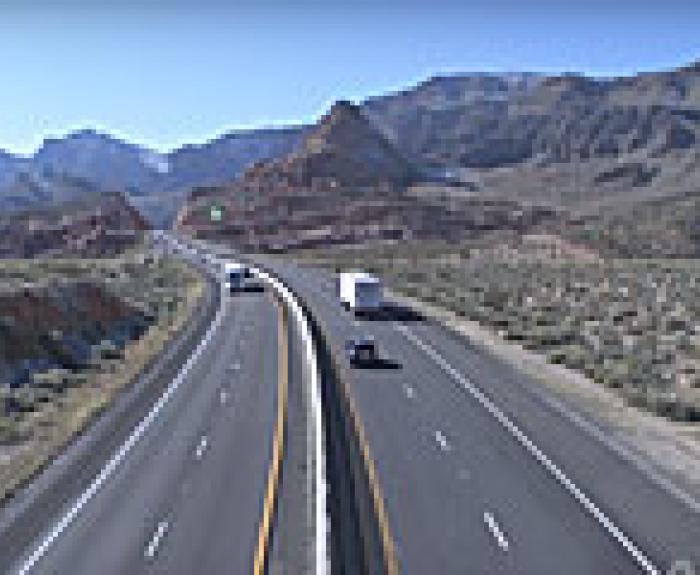 Cars and a truck drive on a divided highway through a desert landscape with mountains in the distance.