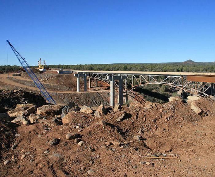 A bridge under construction with a crane, surrounded by dirt and rocks under a clear blue sky.