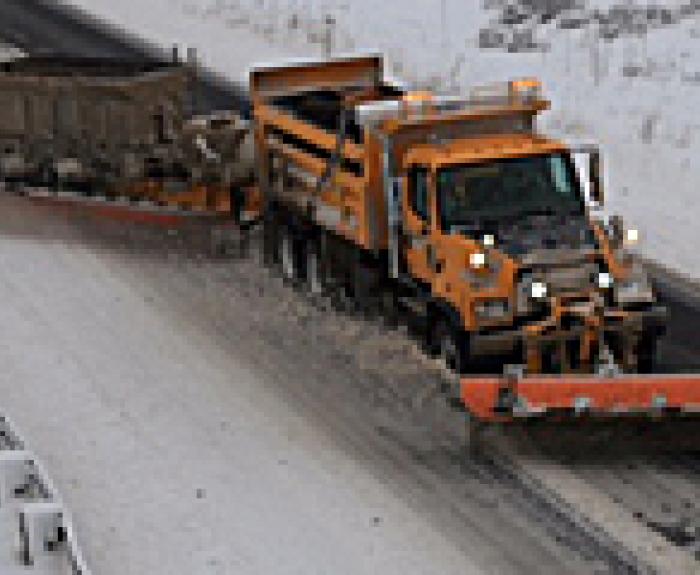 An orange snow plow truck clears snow from a road, with snowy embankments on either side and a salt spreader attached to the back of the truck.