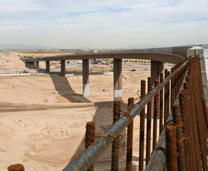 A partially constructed bridge spans a sandy landscape under a cloudy sky, with rusted rebar and parked vehicles visible in the foreground.