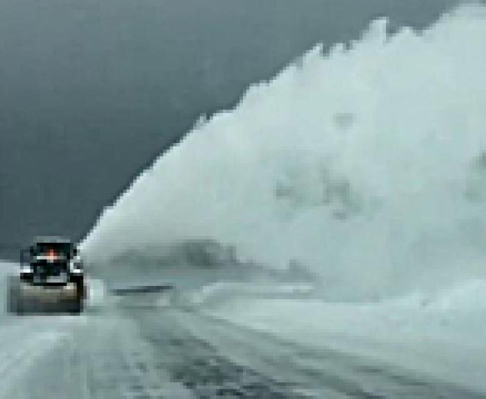 A snowplow clears a snow-covered road, blowing a large plume of snow to the side under a cloudy, gray sky.