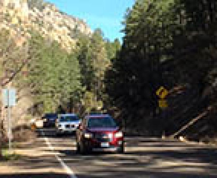 Several cars drive on a winding mountain road surrounded by trees and rocky hillsides under a clear blue sky.