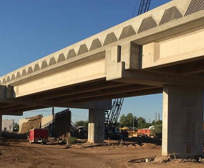 A concrete bridge under construction with support pillars, construction equipment, and dirt visible beneath it on a clear day.