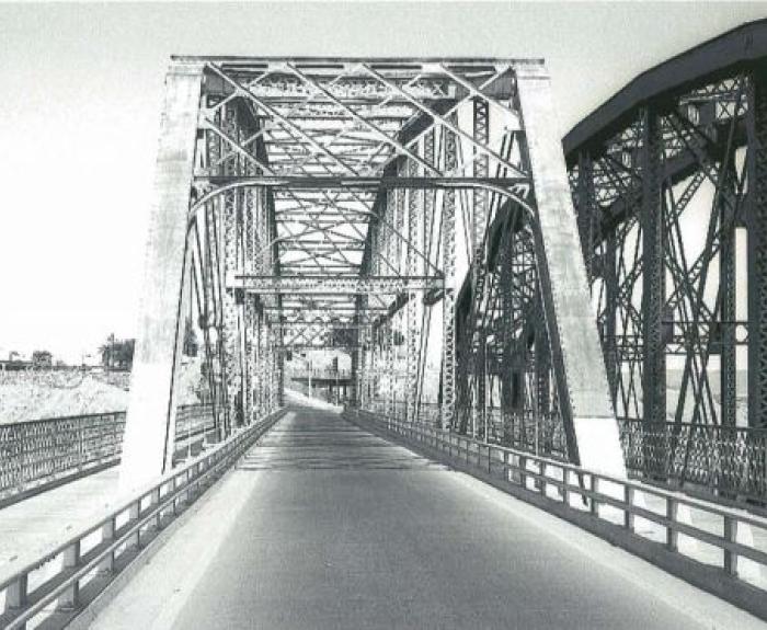 Black and white photo of an old steel truss bridge with an empty roadway, intricate lattice railings, and another similar bridge visible to the right.