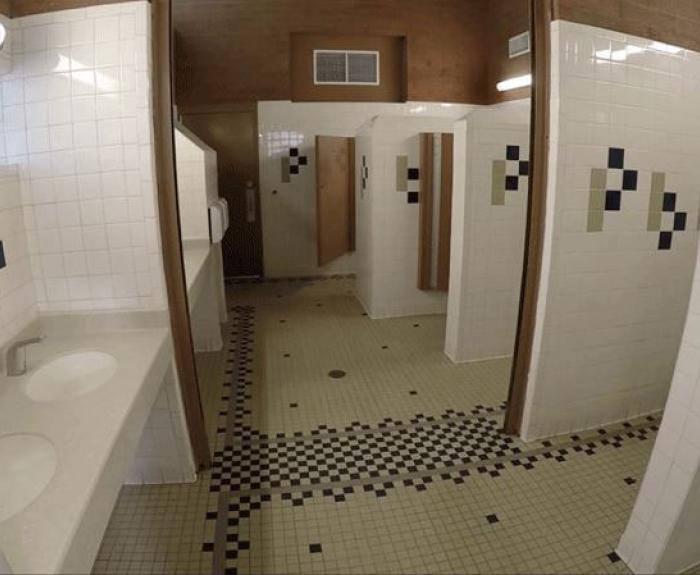 Public restroom with tiled walls and floors, two sinks with a soap dispenser, and several open stalls separated by partitions.