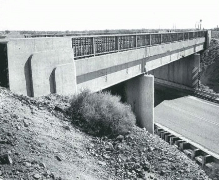 Black and white photo of a concrete bridge with metal railings crossing over a road, with rocky terrain and sparse vegetation surrounding the structure.