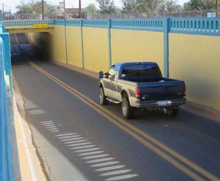 A black pickup truck drives down a two-lane road entering a yellow and blue-painted underpass with a clearance sign of 14 ft 6 in.