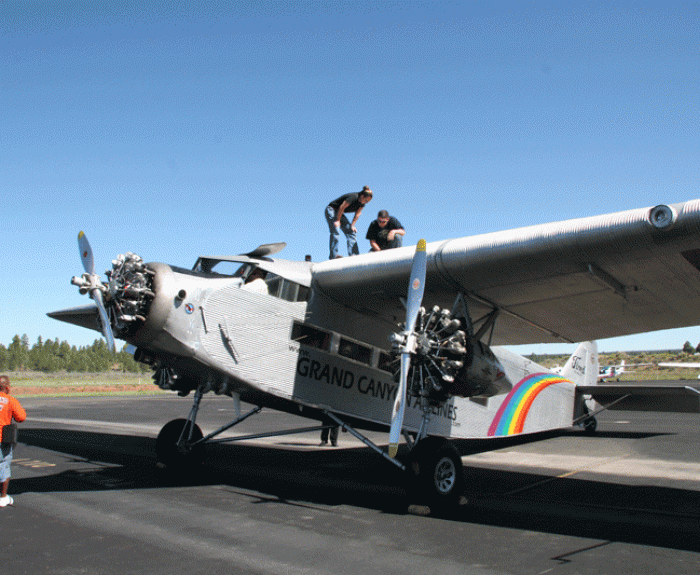 Two people work on the wing of a vintage Grand Canyon airplane parked on an airstrip under a clear sky.