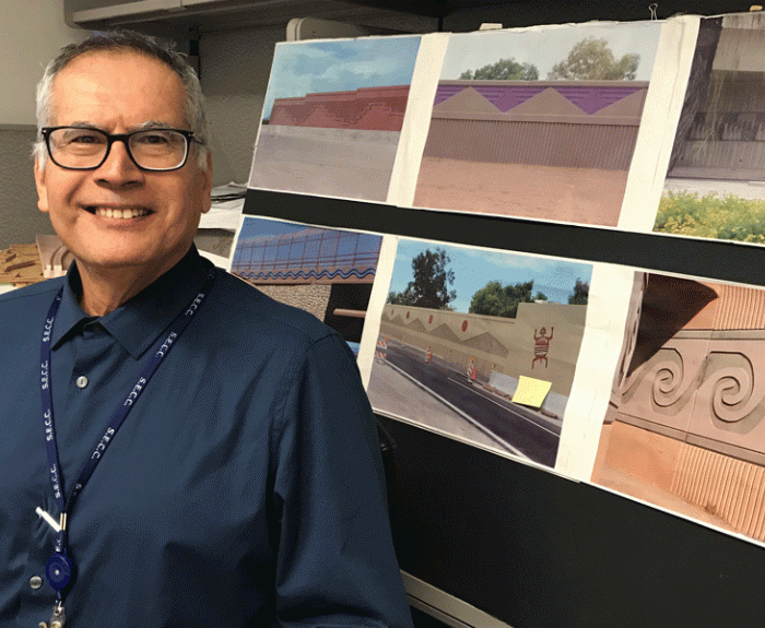 A man in glasses and a blue shirt stands next to a display of printed photos showing decorative wall designs and murals.