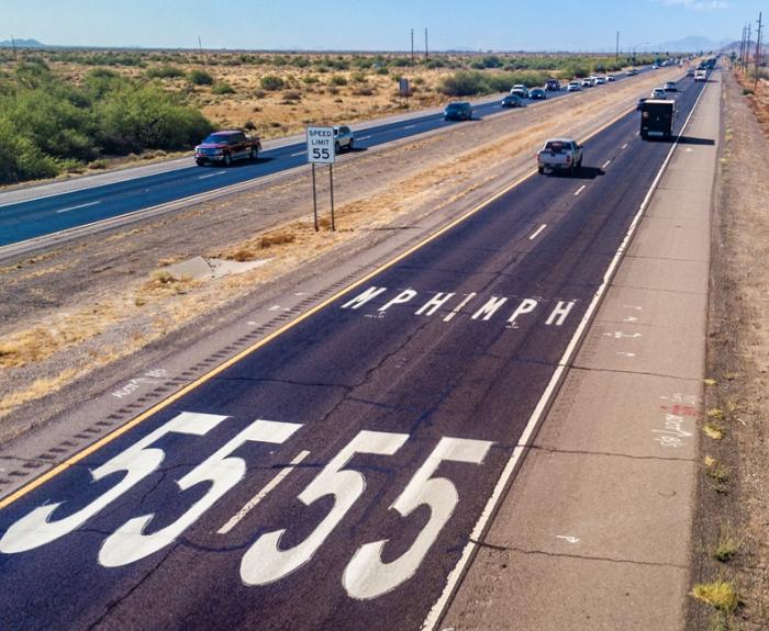 A desert highway with a speed limit of 55 mph painted in large white numbers on the road and several cars driving in both directions.