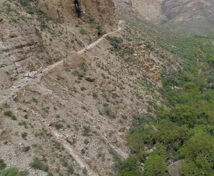 Narrow hiking trail winding along a steep, rocky canyon wall above a green valley.