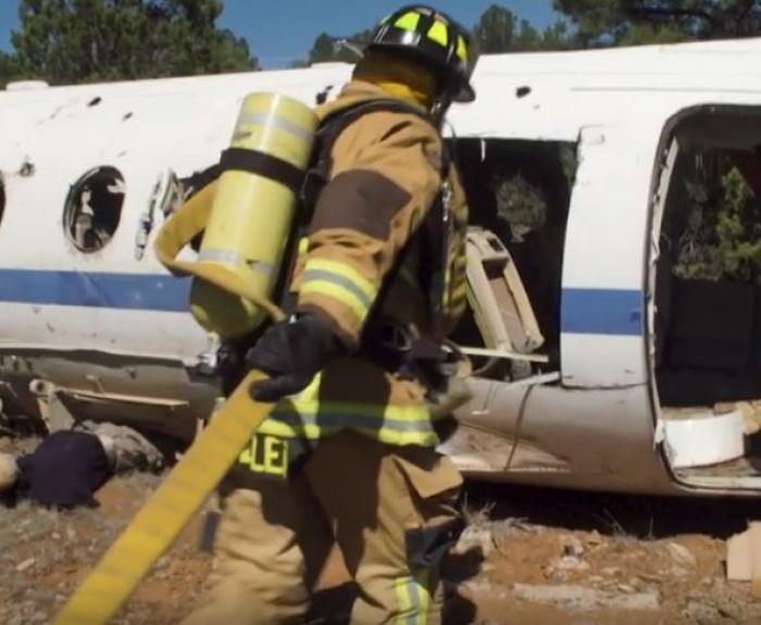 A firefighter in full gear carries a hose near the wreckage of a white airplane, with another person crouched nearby on rocky ground.