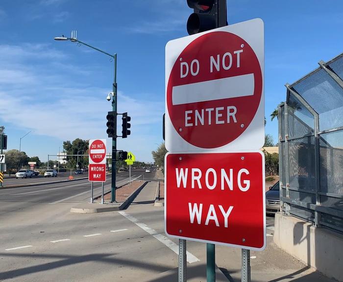 Red Do Not Enter and Wrong Way signs are posted at the entrance to a roadway, with traffic lights and a fence visible on the right side.