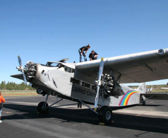 Two people stand on the wing of a silver vintage airplane labeled “Grand Canyon Airlines” with rainbow stripes, while others stand nearby on the tarmac.
