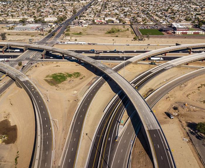 Aerial view of a large highway interchange with multiple overpasses crossing, surrounded by roads, dirt areas, and a residential neighborhood in the background.