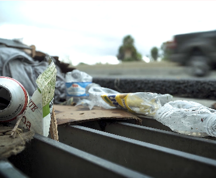 Litter including plastic bottles, a can, and cardboard are scattered on a storm drain by a roadside. A blurred truck drives past in the background.