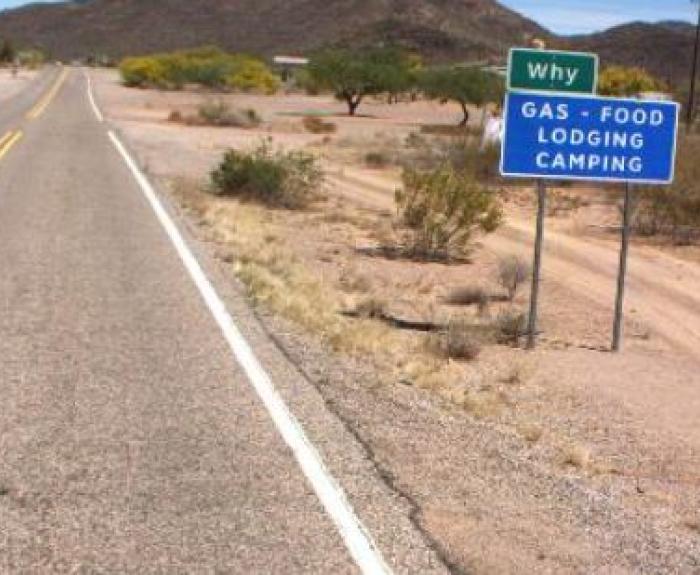 A blue roadside sign reads, Why: Gas, Food, Lodging, Camping beside a deserted highway in a dry, desert landscape with distant hills.