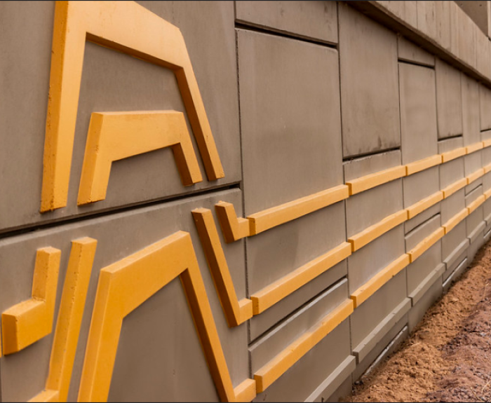 A concrete retaining wall features angular yellow geometric patterns; the wall extends into the distance with gravel at its base and a cloudy sky above.