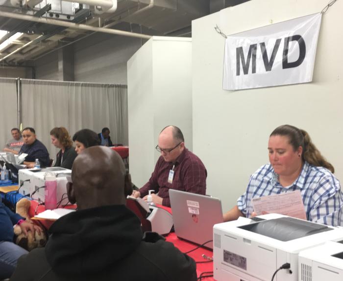 People seated at a DMV counter, staff assisting customers with paperwork and computer tasks.