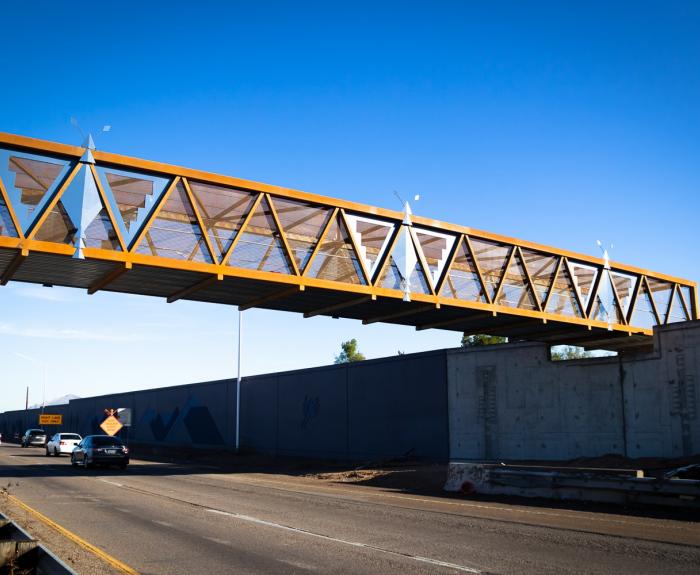 A modern pedestrian bridge with a geometric design spans over a busy highway under a clear blue sky. Cars travel beneath the bridge.