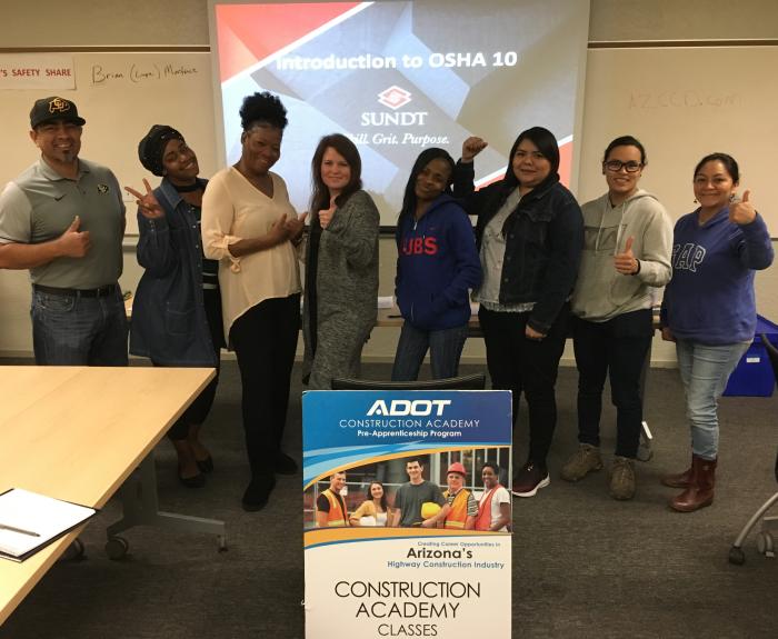 Eight people stand smiling and giving thumbs up in a classroom, with a sign for ADOT Construction Academy Classes and an OSHA 10 presentation on the screen behind them.