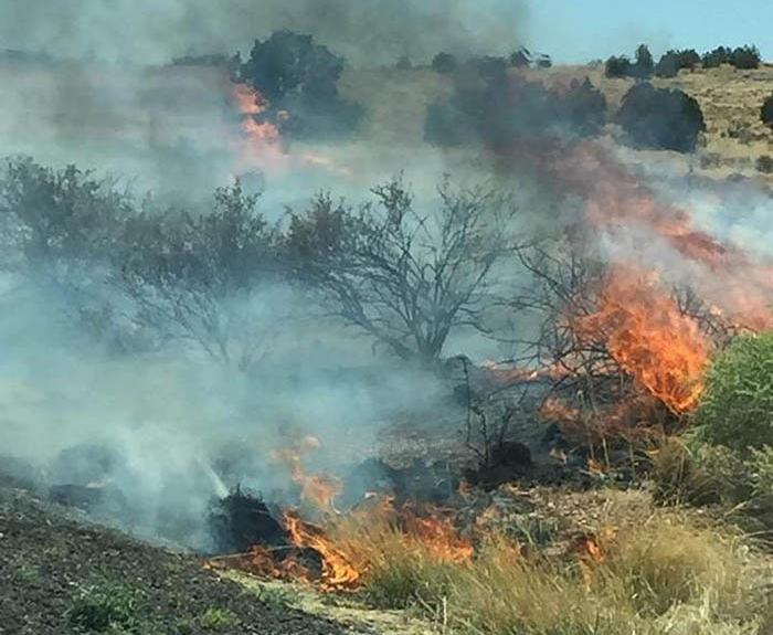 Brush fire burning through dry grass and shrubs in a rural area, producing thick smoke and orange flames under a partly cloudy sky.