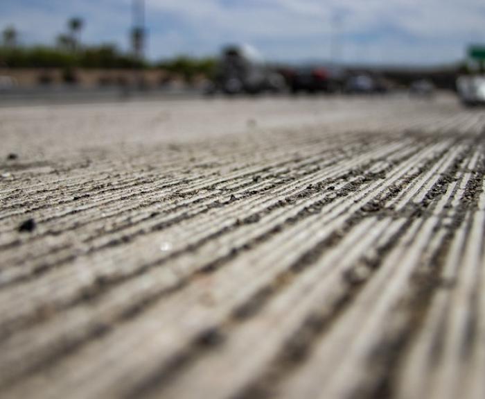 Close-up view of textured grooves on a concrete road surface, with blurred vehicles and highway scenery in the background under a partly cloudy sky.
