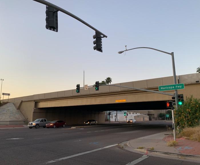 Intersection at dusk with traffic lights, cars passing, and a freeway overpass. A green sign reads Maricopa Fwy. Some palm trees and plants are visible.