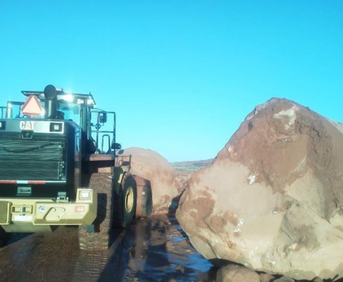 A large bulldozer faces two massive boulders blocking a dirt road under a clear blue sky. The boulders appear to have fallen onto the road, obstructing passage.