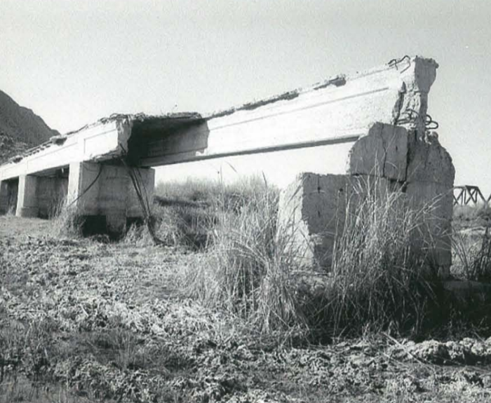 A broken concrete bridge with missing sections stands over dry, cracked ground and tall grass, with a hill and another bridge visible in the background.