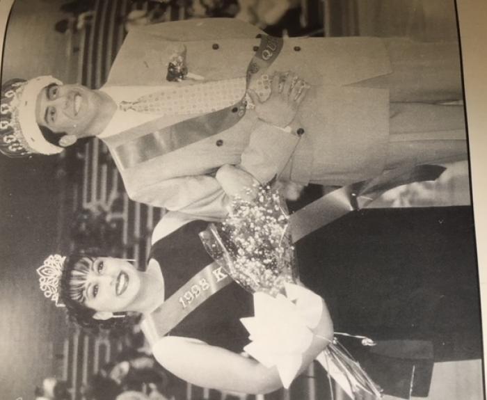 A woman and a man, both wearing sashes and crowns, stand side by side in a gymnasium. The woman holds a bouquet and both are smiling. The sashes read 1992 and King.
