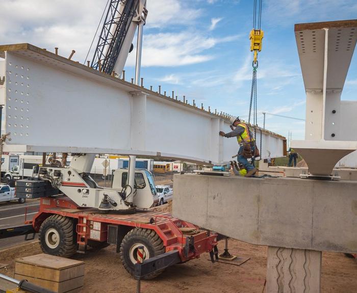 A construction worker in safety gear secures a large steel beam with bolts on a bridge structure while a crane and other equipment are visible nearby.