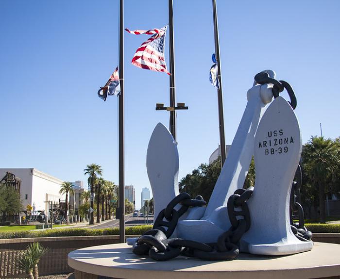 Large anchor monument with USS Arizona BB-39 inscribed, American flags flying behind, palm trees in background.