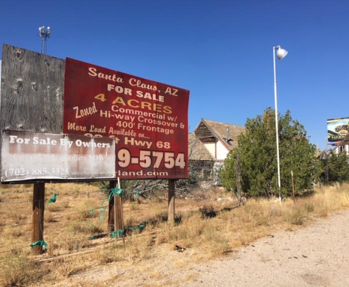 Large red and white billboard advertising land for sale in Santa Claus, AZ, next to an old building and overgrown weeds on a sunny day.
