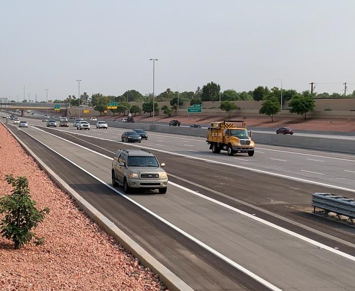 A multi-lane highway with several cars and a yellow utility truck driving under clear skies, bordered by red gravel landscaping and small trees.