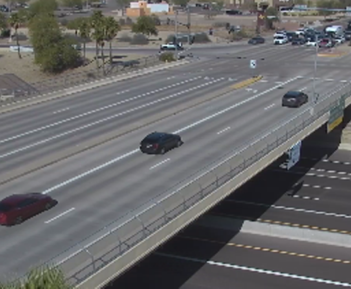 An overpass crosses above a multi-lane road with light traffic; several cars travel on both the overpass and the street below, with trees and buildings in the background.