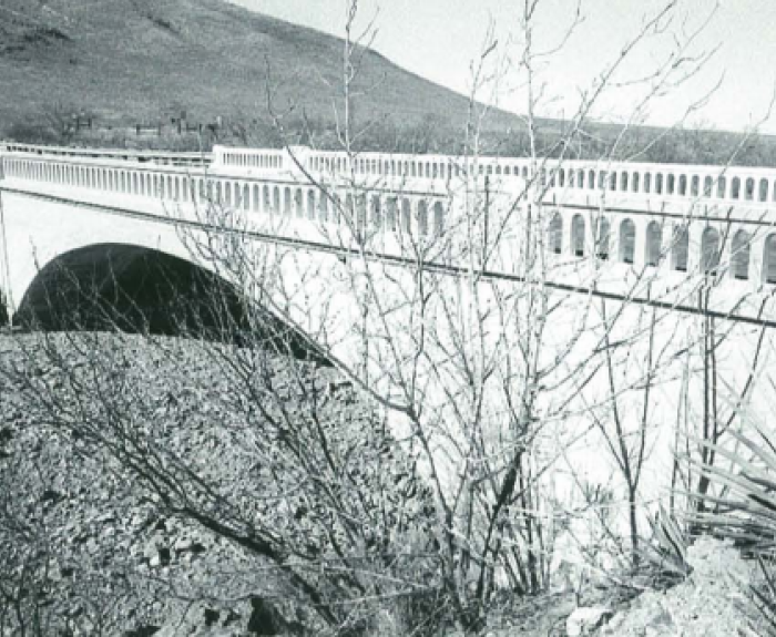 Black and white photo of a concrete bridge with arched railings spanning a dry, rocky creek bed, with sparse vegetation and a hill in the background.