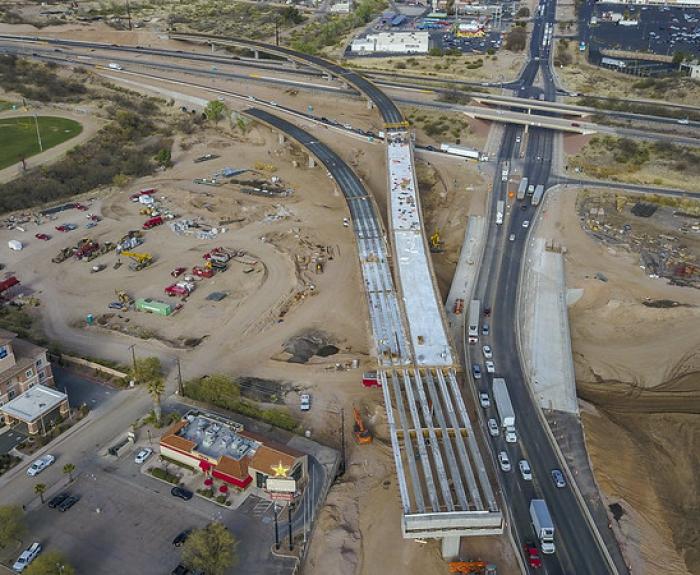 Aerial view of a highway under construction with vehicles, equipment, and buildings nearby; part of the road is unfinished while traffic moves on adjacent lanes.