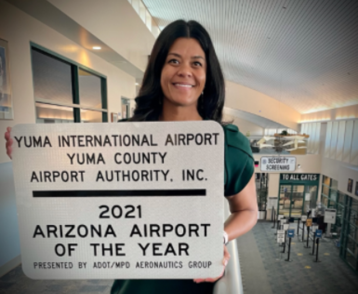 A woman stands indoors holding a sign that reads Yuma International Airport, 2021 Arizona Airport of the Year, with airport gates and security in the background.