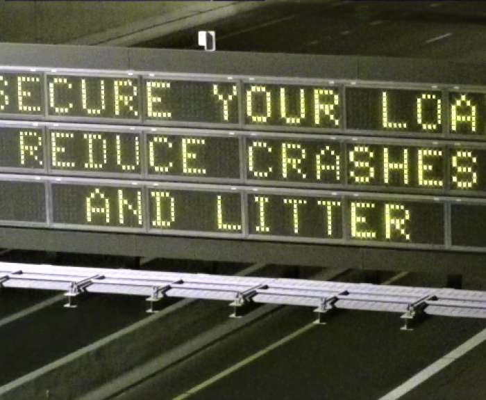 Overhead digital highway sign reads SECURE YOUR LOAD REDUCE CRASHES AND LITTER.