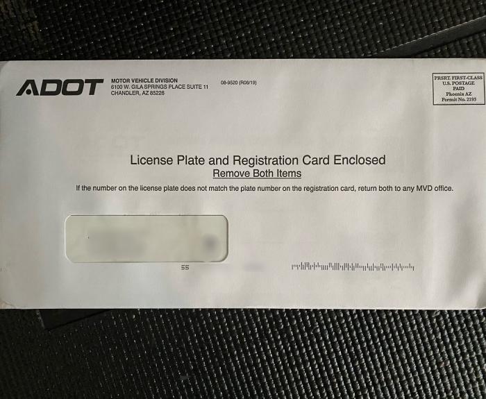 A white envelope from ADOT Motor Vehicle Division with a window showing an address, marked License Plate and Registration Card Enclosed on a black textured surface.