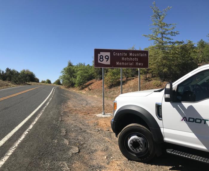 A white ADOT truck is parked on the roadside near a brown sign marking State Route 89 as Granite Mountain Hotshots Memorial Hwy, under a clear sky.