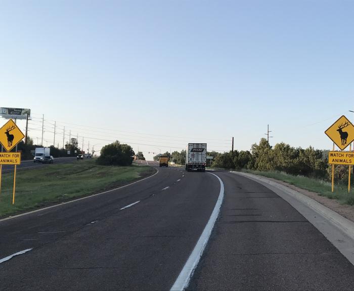 A highway with two yellow animal crossing signs reading Watch for Animals and a truck driving ahead under a clear sky. Trees line both sides of the road.