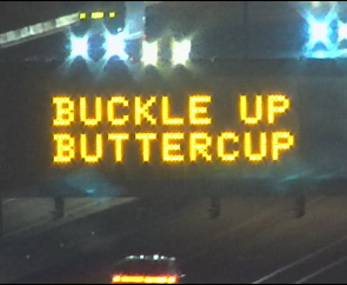 Electronic highway sign at night displays the message BUCKLE UP BUTTERCUP in bright yellow text above lanes of traffic.