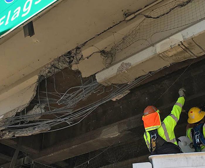 Two construction workers in safety gear inspect and repair a damaged overpass with exposed rebar and cracked concrete beneath a green highway sign.