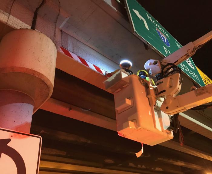 A worker in a bucket places reflective tape on the underside of a bridge deck. The scene is illuminated by work lights.