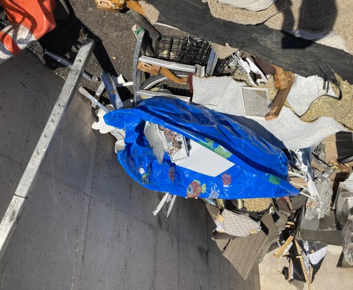 A blue inflatable kiddie pool decorated with with cartoon fish designs sits among various discarded household items in a dumpster under bright sunlight.