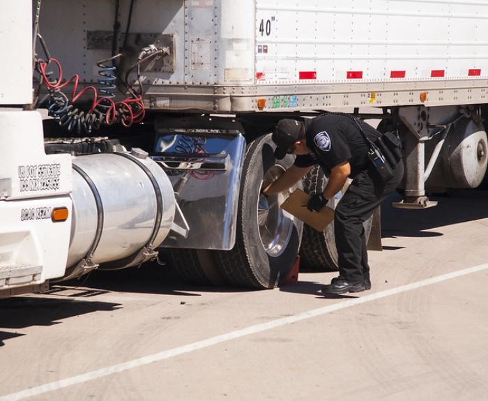 A police officer inspects the tires of a parked semi-truck on a sunny day.