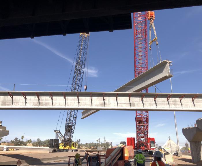 A large crane lifts a concrete bridge beam into place at a construction site, with workers in safety gear observing below.
