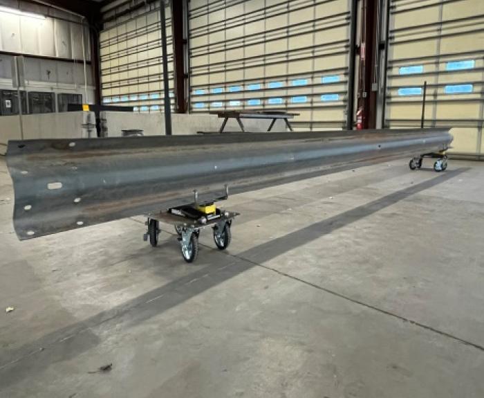 A long metal guardrail rests on two rolling carts inside a spacious industrial warehouse with large doors and concrete floors.